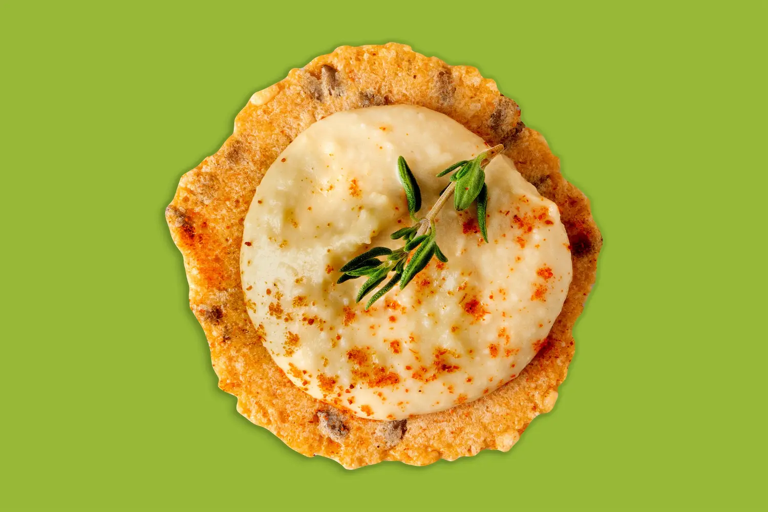 Round cracker topped with creamy dip and a thyme sprig on a green background.