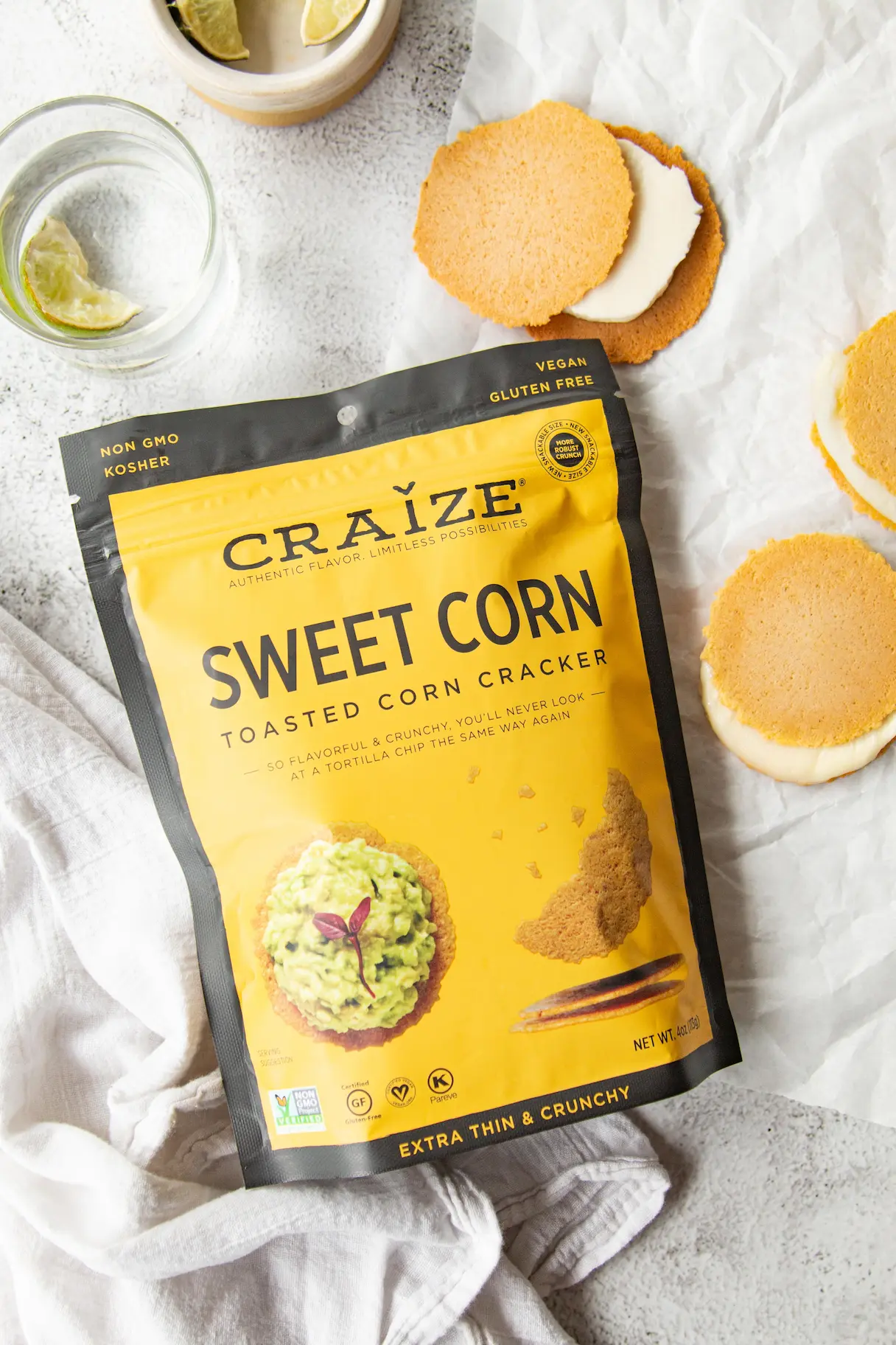 Craize Sweet Corn crackers bag on linen with alfajores, lime wedges and a glass of water.