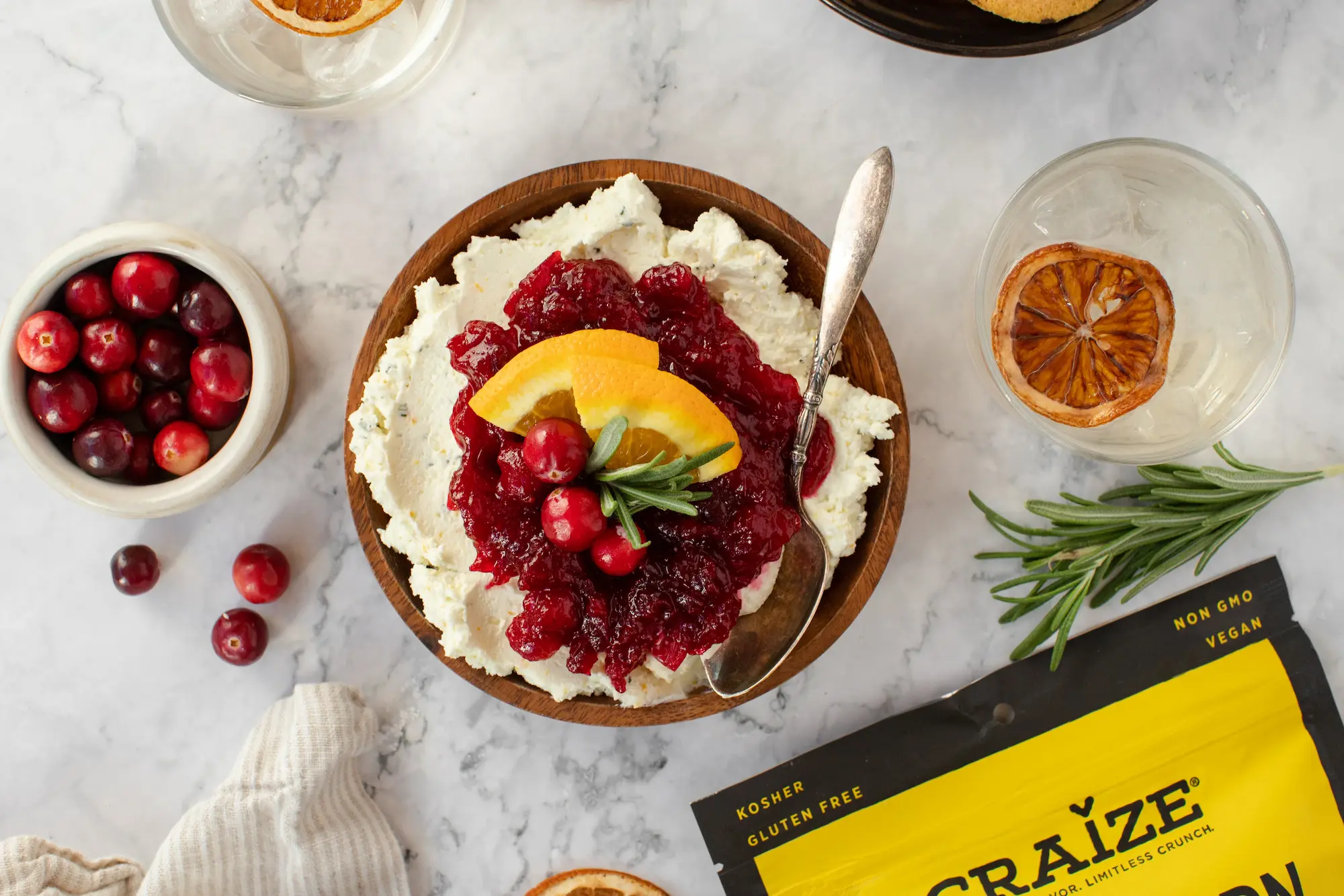 Bowl of creamy dip topped with cranberry sauce, orange slices and rosemary on a marble surface.