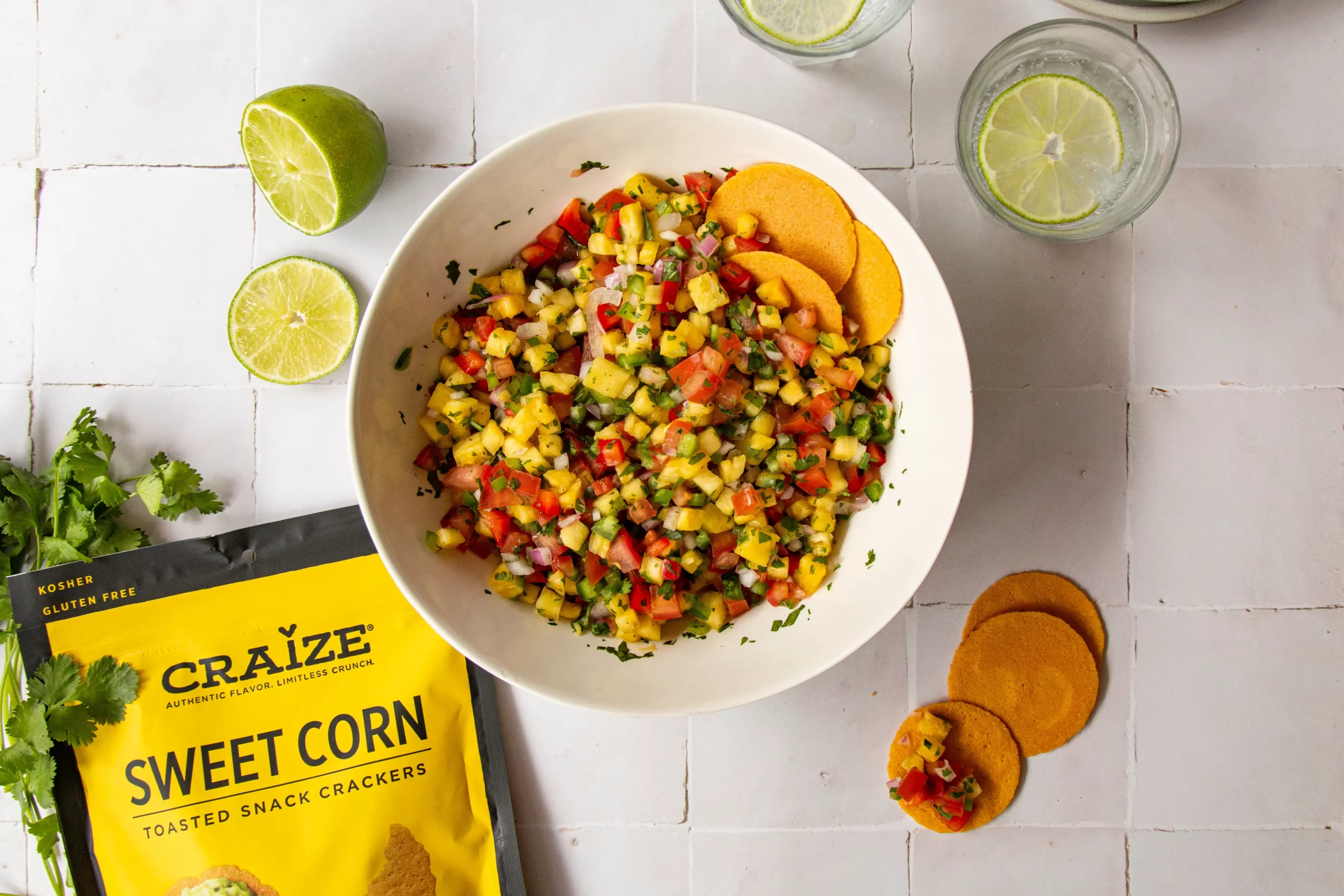 Bowl of chopped salsa with corn crackers, limes, and a Craize Sweet Corn crackers bag on a white tabletop.